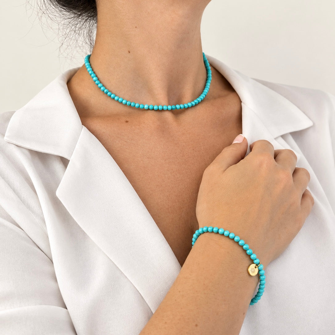 A close-up of a model wearing a turquoise beaded choker necklace and a matching turquoise beaded bracelet with a small gold disc charm, styled with a white collared shirt.