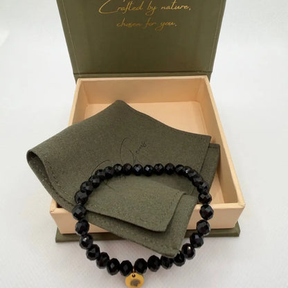 Black Tourmaline Faceted Bracelet with gold charm on an olive green pouch in a cream and olive green branded box and light background