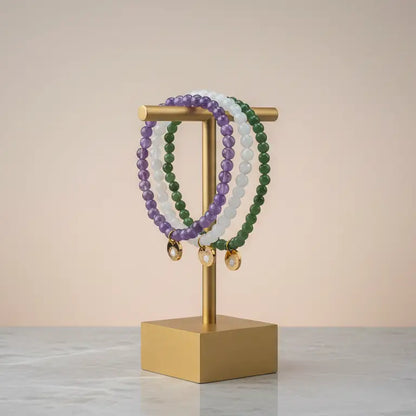 Amethyst, Moonstone, and Nephrite Jade beaded bracelets with gold charms on a metallic geometric stand and neutral background