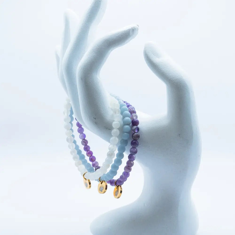 Amethyst, Angelite, and Moonstone beaded bracelets with gold charms on a white mannequin hand against a light blue background