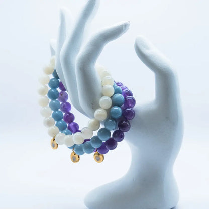 Amethyst, Angelite, and Moonstone beaded bracelets with gold charms on a white mannequin hand against a light background