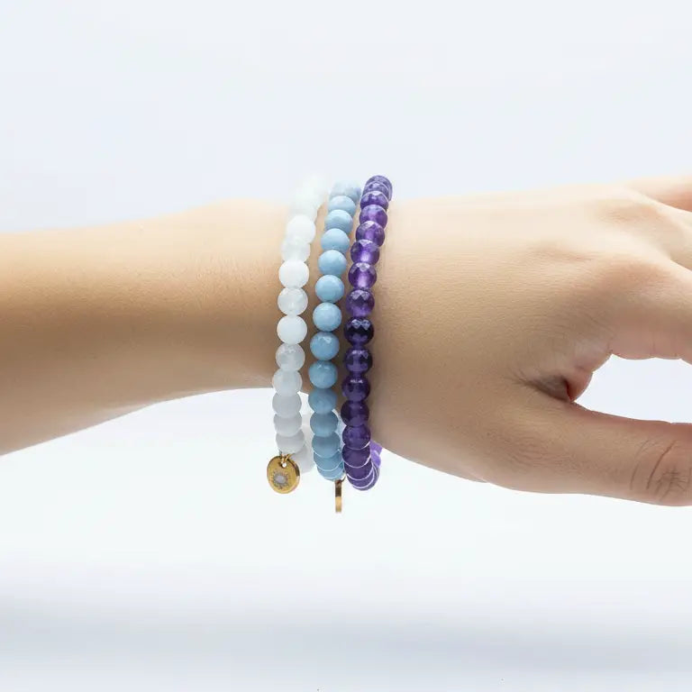 Hand wearing Amethyst, Angelite, and Moonstone beaded bracelets with gold charms on a light background