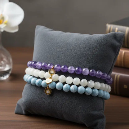 Stack of Amethyst, Angelite, and Moonstone beaded bracelets with gold charms on a gray pillow with books and a flower in the background