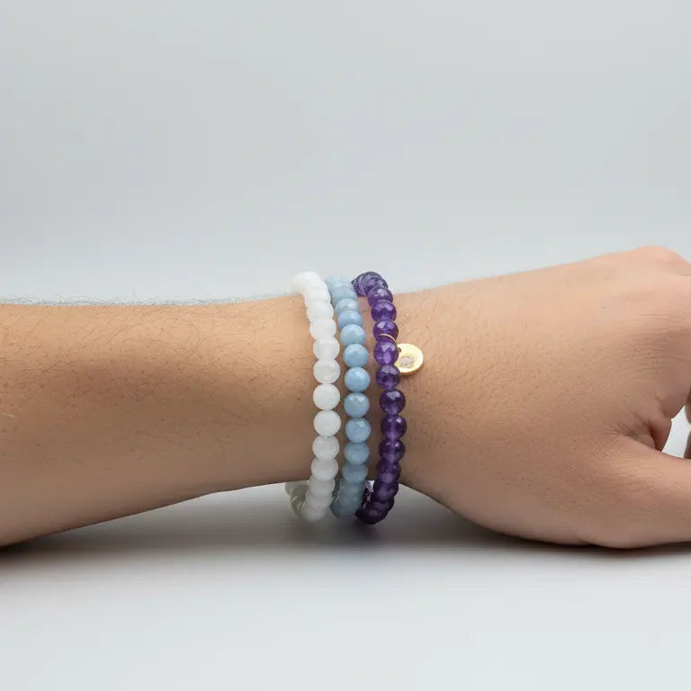 Hand wearing Amethyst, Angelite, and Moonstone beaded bracelets with gold charm on a plain background