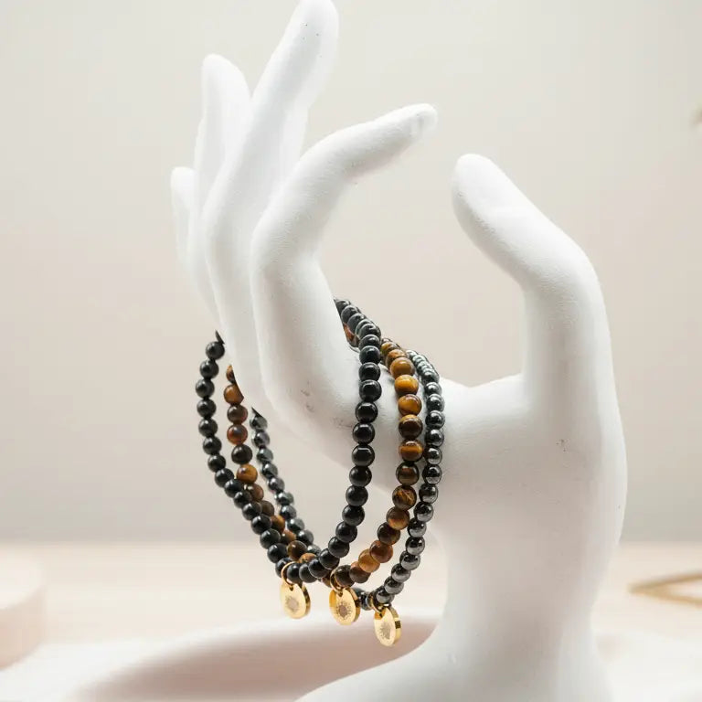 Mannequin hand with Tiger Eye, Hematite, and Obsidian beaded bracelets and gold charms on a blurred neutral background