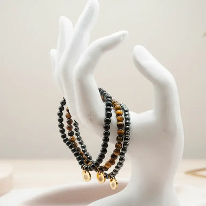 Mannequin hand with Tiger Eye, Hematite, and Obsidian beaded bracelets and gold charms on a blurred neutral background