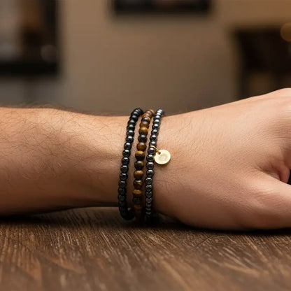 Wrist with Tiger Eye, Hematite, and Obsidian beaded bracelet with gold charm on a wooden surface and blurred indoor background