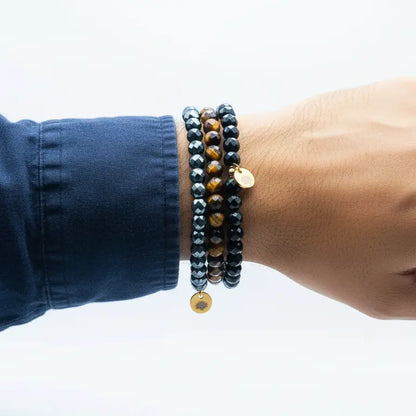 Tiger Eye, Hematite, and Obsidian faceted beaded bracelets with gold charms on a wrist with nany sleeve and neutral background