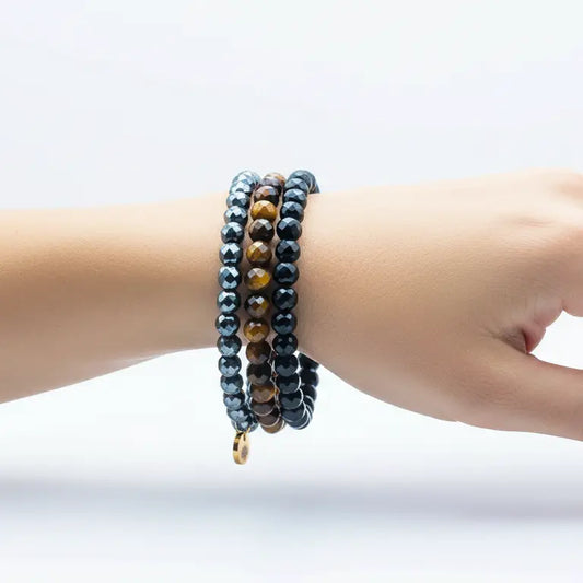 Tiger Eye, Hematite, and Obsidian faceted beaded bracelets with gold charm on a wrist and light grey background.