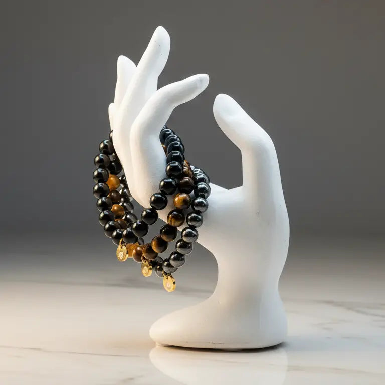 Ceramic hand-like stand with Tiger Eye, Hematite, and Obsidian beaded bracelets with gold charms on a marble top and grey background