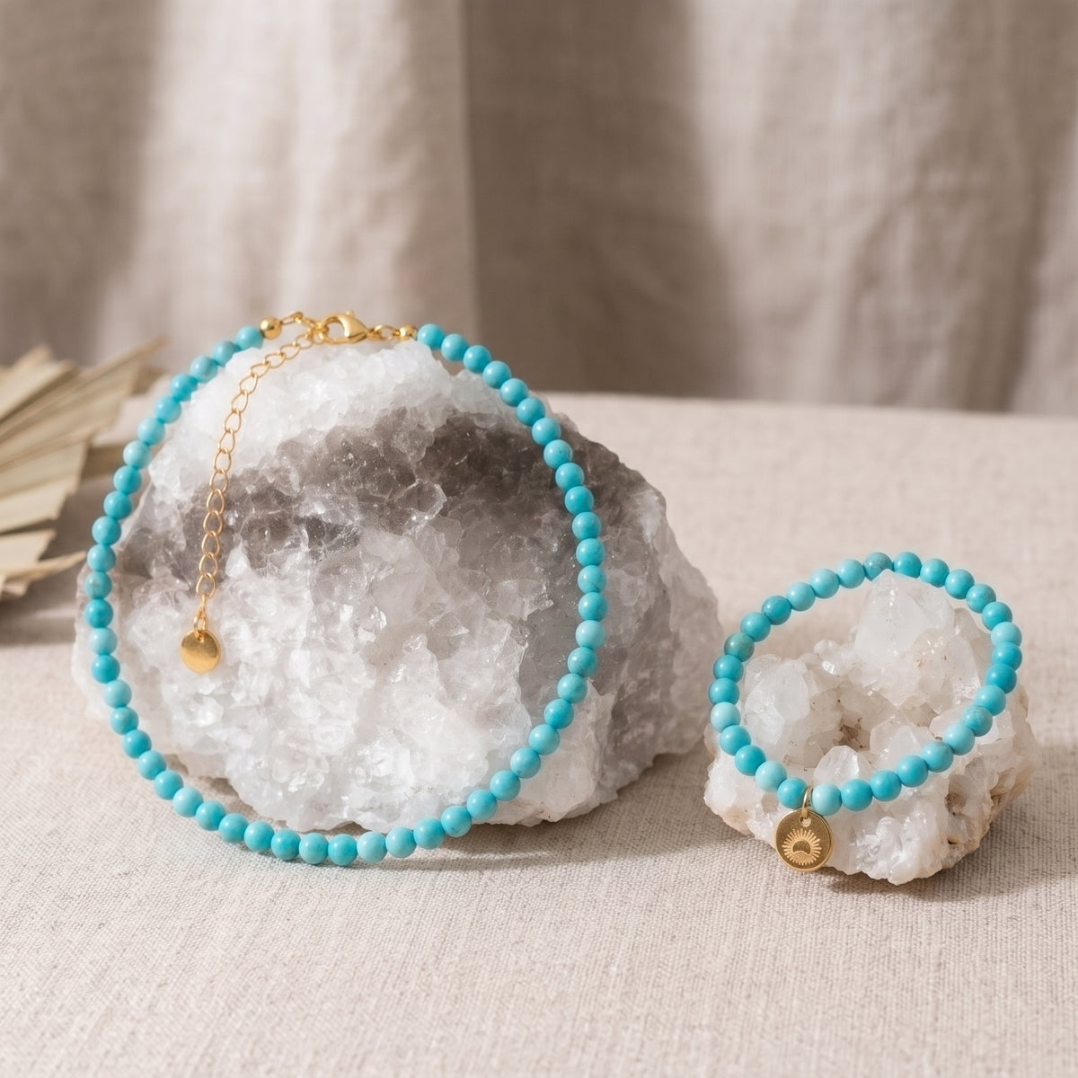 A turquoise beaded necklace and matching bracelet displayed on white crystal geodes, with a gold chain extender and small gold bell charm on a neutral fabric background.