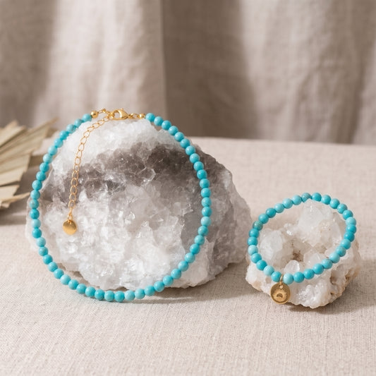A turquoise beaded necklace and matching bracelet displayed on white crystal geodes, with a gold chain extender and small gold bell charm on a neutral fabric background.