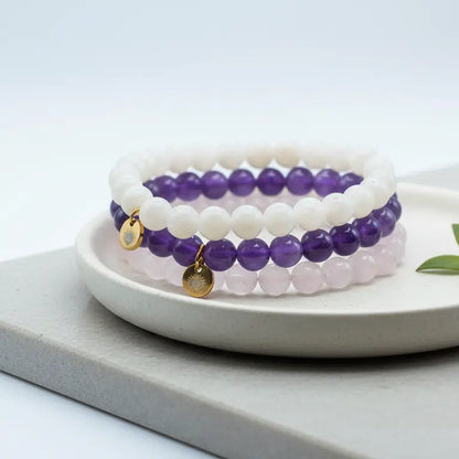Rose Quartz, Moonstone, and Amethyst beaded bracelets with gold charms on a white plate with a light gray background