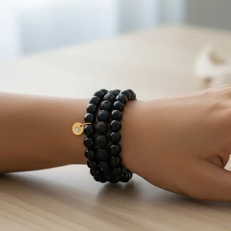 Hand wearing Black Tourmaline, Obsidian, and Lava Stone \beaded bracelets with gold charm on a wooden surface and blurred background