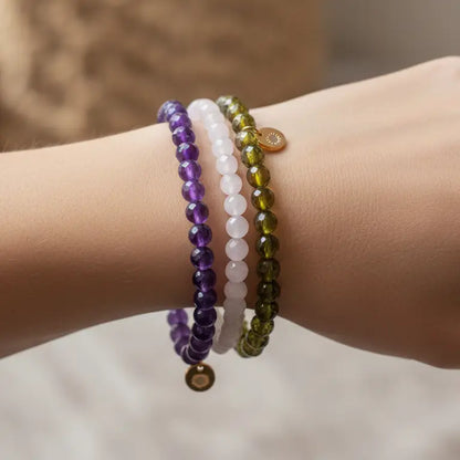 Close-up of a wrist wearing Rose Quartz, Amethyst, and Peridot faceted beaded bracelets with gold charms on a blurred background
