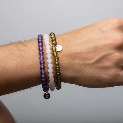 Hand wearing Rose Quartz, Amethyst, and Peridot faceted beaded bracelets with gold charms on a gray background