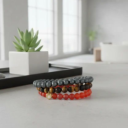 Carnelian, Tiger Eye, and Hematite beaded bracelets with gold charm on a white surface with a flower pot and blurred indoor background