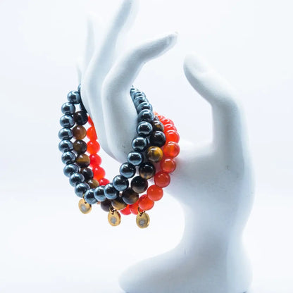 Stack of Carnelian, Tiger Eye, and Hematite beaded bracelets with gold charms on a white mannequin hand against a white background