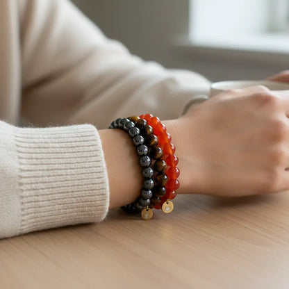 Person wearing Carnelian, Tiger Eye, and Hematite beaded bracelets with gold charms on a wooden table
