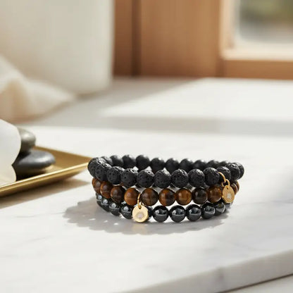 Hematite, Tiger Eye, and Lava Stone beaded bracelets with gold charms on a marble surface with a decorative plate of stones and window in the background.