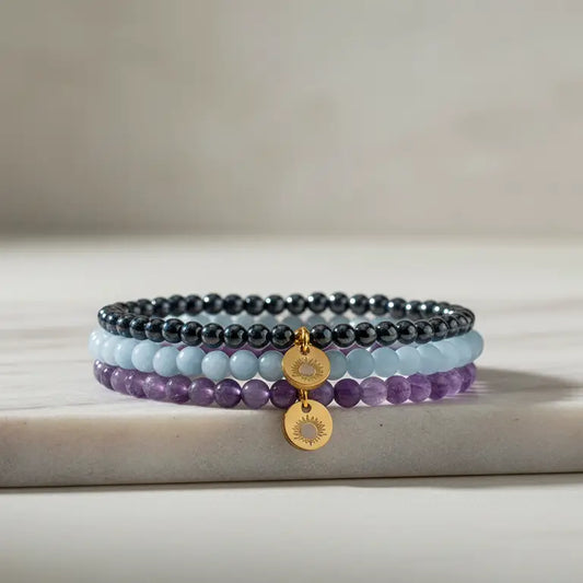 A set of Amethyst, Angelite, and Hematite beaded bracelets with gold charms on a marble sand with light grey background