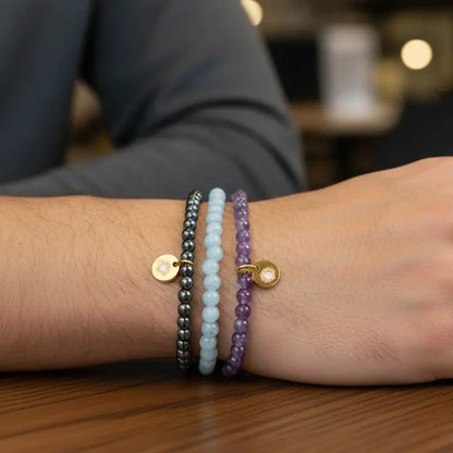 Amethyst, Angelite, and Hematite beaded bracelets with gold charms on a wrist against a wooden stand with grey background