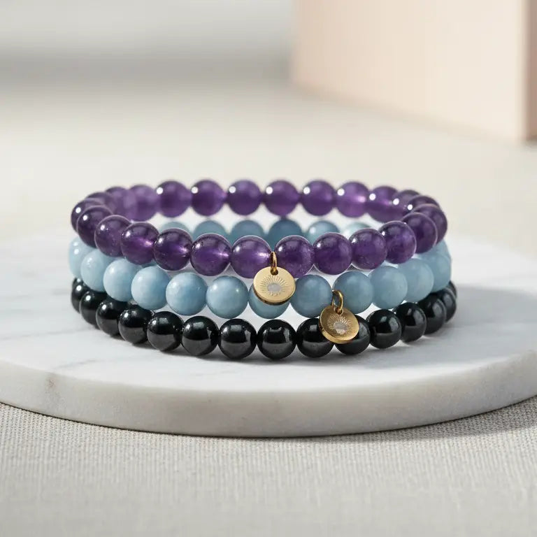 A set of Amethyst, Angelite, and Hematite beaded bracelets with gold charms on a marble stand and light background