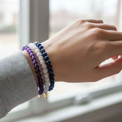 Hand wearing Amethyst, Moonstone, and Lapis Lazuli faceted beaded bracelets with gold charm by a window