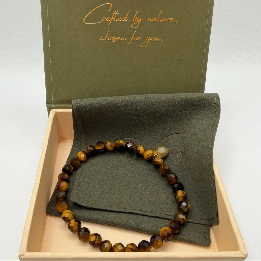 Tiger Eye Faceted Bracelet with gold charm against an olive green pouch in a cream and olive green branded box and light grey background
