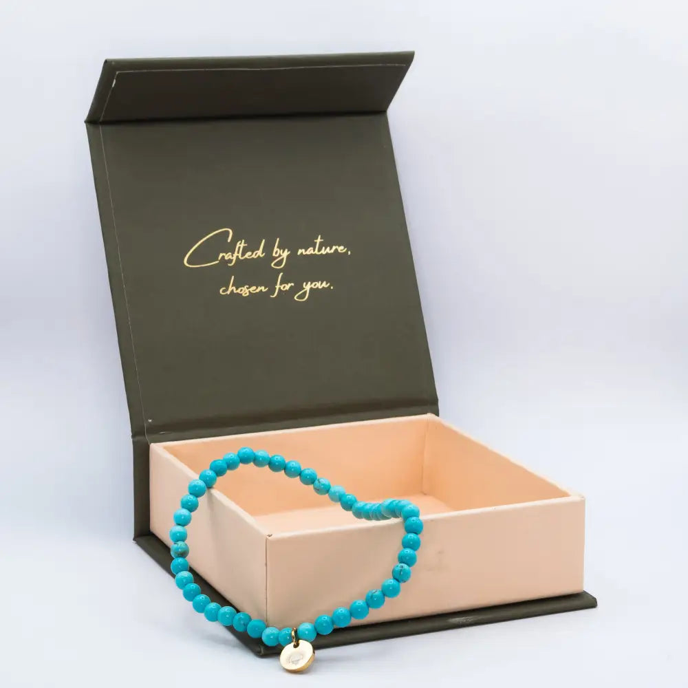 Turquoise Beaded Bracelet with gold charm on a cream and olive green branded box and light grey background