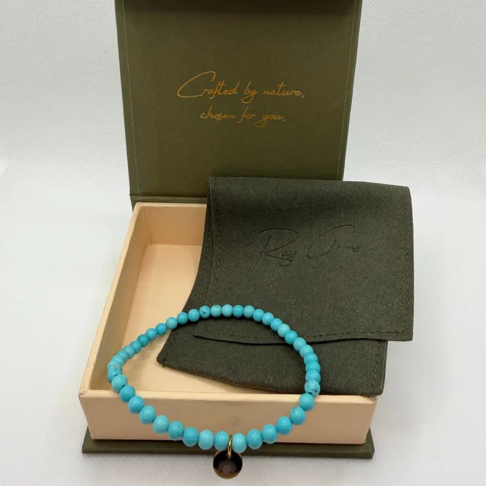 Turquoise Beaded Bracelet with gold charm next to an olive green pouch in a  cream and olive green branded box and netural background