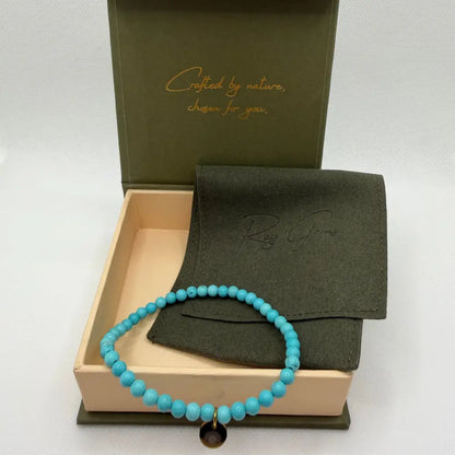 Turquoise Beaded Bracelet with gold charm next to an olive green pouch in a  cream and olive green branded box and netural background